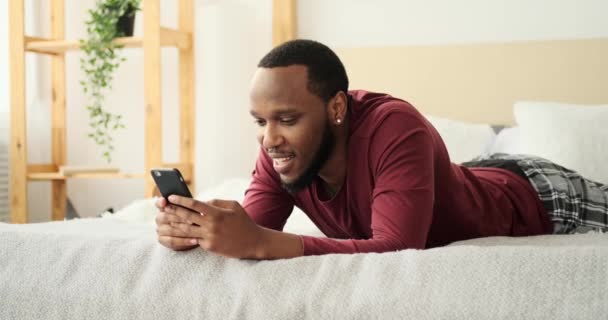 Afroamerikaner liegt auf Bett und benutzt Handy — Stockvideo