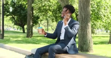 İş adamı kahve içiyor ve şehir parkında cep telefonuyla konuşuyor.