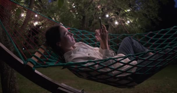 Egy nő mobiltelefont használ, miközben éjjel függőágyon fekszik. — Stock videók
