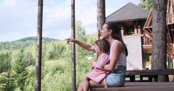 Женщина показывает пальцем, разговаривая со своей дочерью, сидящей на деревянной скамейке на природе — стоковое видео