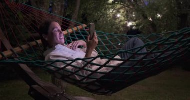 Geceleri hamakta uzanırken cep telefonu kullanan bir kadın görüntüsü.