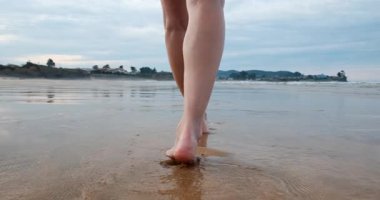 Yetişkin bir kadının çıplak ayaklarıyla okyanusa doğru yansıyan ıslak kumda bulutlu mavi gökyüzünün altında ağır çekimde yürümesini yakından görmek..