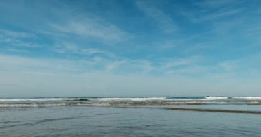 Sabit geniş deniz manzarası yavaş çekimde küçük dalgalar mavi bir gökyüzünün altında ıslak kumun üzerinde yuvarlanıyor..