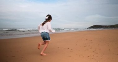 Bir kız çocuğu ıslak kıyı şeridi boyunca ağır çekimde koşar. Arka planda dalgalar bulutlu mavi bir gökyüzünün altında..
