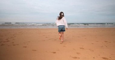 Bir kız çocuğu kumlu bir sahilde okyanusa doğru ağır çekimde koşuyor, bulutlu mavi bir gökyüzünün altında ıslak kumların üzerinde ayak izleri bırakıyor..
