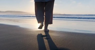 Islak kumda ve yumuşak dalgaların içinde çıplak ayakla yürüyen bir kadının yavaş çekim görüntüsü. Altın güneş kıyı şeridine yansıyor.