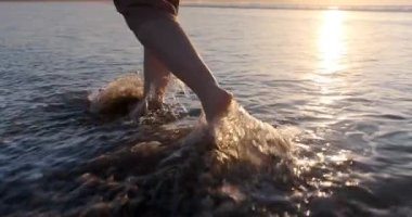 Yavaş çekim, çıplak ayaklı bir kadının kumdan sığ okyanus dalgalarına doğru yürümesi, altın saatinde suyu sıçratması..