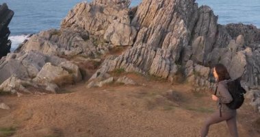 Sırt çantalı genç bir kadın dramatik bir kayalığın kenarına yürür ve okyanus dalgalarına bakmak için durur..