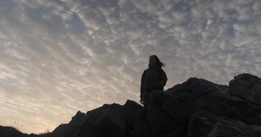 Sırt çantasıyla kayalık bir zirveye tırmanan, dramatik bulutlu bir gökyüzüne karşı silueti olan bir kadının düşük açılı görüntüsü..