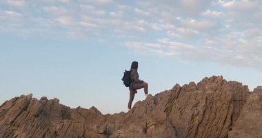 Sırt çantalı genç bir kadın yürüyüşçünün yavaş çekimde kayalık bir tepede dikilip bulutlu mavi gökyüzüne bakarken..