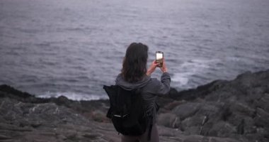 Sırt çantalı genç bir kadının kayalık kayalıklarda dikilirken telefonuyla dalgalı denizin fotoğraflarını çekerken görüntüsü..