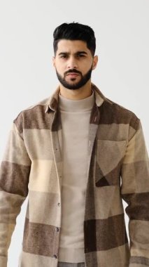 Genç yetişkin bir adam beyaz bir arkaplan karşısında duruyor, kameraya ciddi ciddi bakıyor ve onaylamadığı ya da reddettiği için parmağını sallıyor..