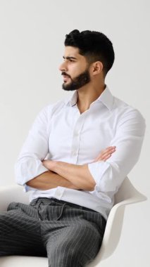 Kolları beyaz bir arka planda çapraz bir sandalyede oturan sakallı, yakışıklı, genç bir adamın portresi. Güvenle gülümsüyor..