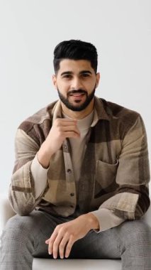 Sakallı, yakışıklı ve şık genç bir adamın portresi, beyaz bir arka plana karşı bir sandalyede oturuyor ve kameraya gülümsüyor..