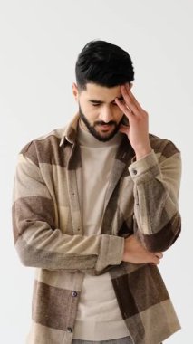 Genç yetişkin bir adam beyaz bir arka plana karşı duruyor, stresli ve endişeli görünüyor. Kafasını elinde tutuyor, baş ağrısı, endişe ya da tükenme belirtileri gösteriyor..