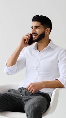 Gülümseyen genç bir iş adamı akıllı telefonuyla dostça ve olumlu bir konuşma yapar. Beyaz bir arka plana karşı sandalyede otururken el kol hareketi yapar..