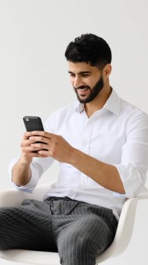 Genç, yetişkin bir iş adamı beyaz bir arkaplana karşı sandalyede oturuyor, akıllı telefonuyla yazarken gülümsüyor ve selfie çekiyor..