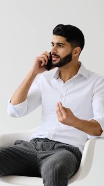 Genç bir iş adamı akıllı telefonuyla ciddi bir konuşma yapar. Beyaz bir arka plana karşı sandalyede otururken el kol hareketi yapar..