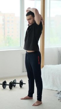 Spor kıyafetleri içinde genç bir adam ısınma egzersizleri yapıyor. Kollarını esnetme ve zıplama hareketleri de dahil. Ev antrenmanından önce yatak odasında..