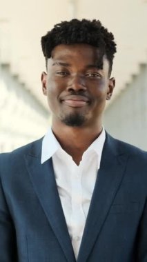 Yavaş çekimde, kendine güvenen ve başarılı genç, takım elbiseli siyahi bir iş adamının modern bir binadaki kameraya gülümseyen portresi..