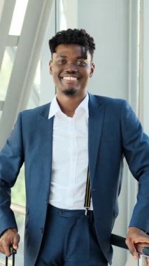 Bir havaalanında ya da modern bir binada bavul taşırken kameraya gülümseyen takım elbiseli, kendine güvenen genç siyahi bir işadamının portresi..