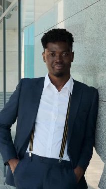 Modern bir binaya yaslanmış ve kameraya gülümseyen takım elbiseli yakışıklı ve şık genç bir siyahi iş adamının yavaş çekim portresi..