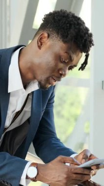 Modern bir bina ya da havaalanı terminalinde beklerken akıllı telefonunu kullanan gülümseyen genç bir siyahi işadamının yavaş çekim yakın çekimi..