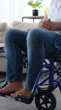 Sakallı, çıplak ayaklı, mutlu bir son sınıf öğrencisi evde tekerlekli sandalyede oturuyor ve kameraya gülümsüyor. Kamera yukarı doğru eğiliyor. Dikey video.