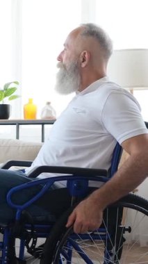 Sakallı mutlu bir son sınıf öğrencisi evde tekerlekli sandalyede oturur, döner ve kameraya sıcak bir şekilde gülümser. Dikey video.