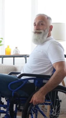 Sakallı mutlu bir son sınıf öğrencisi evde tekerlekli sandalyede oturur, döner ve kameraya sıcak bir şekilde gülümser. Dikey video.