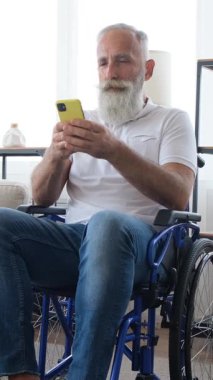 Sakallı, gülümseyen bir son sınıf öğrencisi evde tekerlekli sandalyede oturuyor ve akıllı telefon kullanıyor, göz gezdiriyor ya da mesaj yazıyor. Dikey video.