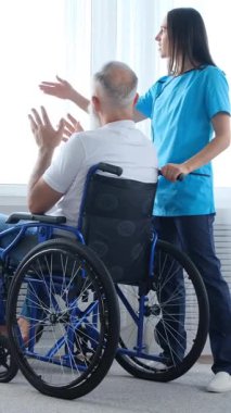 Önlüklü bir kadın hasta bakıcı, tekerlekli sandalyedeki yaşlı bir adamı pencereden aşağı itiyor. Bakımevi ya da evde bakım konsepti. Dikey video.