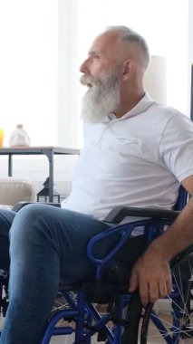 Yaşlı, sakallı, etkileyici bir sakallı adam evde tekerlekli sandalyede oturur, dans eder, müzik eşliğinde şarkı söyler ve güler. Dikey video.
