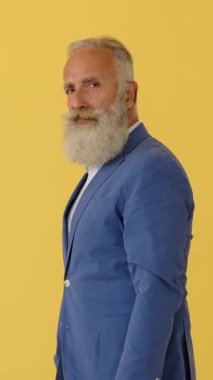 Uzun sakallı, mavi ceketli bir son sınıf öğrencisi kameraya dönüp müstehcen bir orta parmak hareketi gösteriyor. Sarı stüdyo geçmişi. Dikey video.