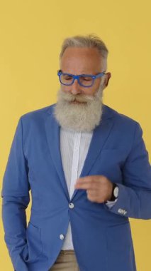 Büyük beyaz sakallı, mavi gözlüklü havalı, şık bir son sınıf öğrencisi parmaklarını şaklatıp dans ediyor. Sarı stüdyo geçmişi. Dikey video.