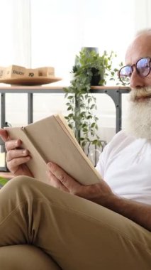 Sakallı ve gözlüklü mutlu bir son sınıf öğrencisi evde oturur, kitap okur ve komik bir hikayeye güler. Dikey video.