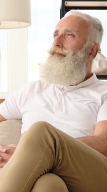 Sakallı düşünceli bir son sınıf öğrencisi evdeki koltukta oturur, gözlerini kaçırır ve düşünceli bir şekilde bıyığını okşar. Dikey video.