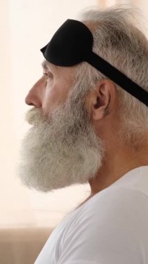 Büyük beyaz sakallı yaşlı bir adam siyah bir uyku maskesi takar ve dinlenmek ya da uyumak için yatağa uzanır. Dikey video.