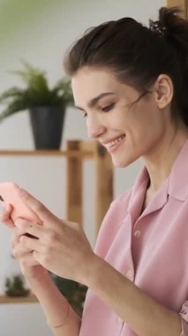 Pembe pijamalı mutlu genç bir kadın evde akıllı telefonuyla sosyal medyaya mesaj atarken ya da gezinirken gülümsüyor. dikey video