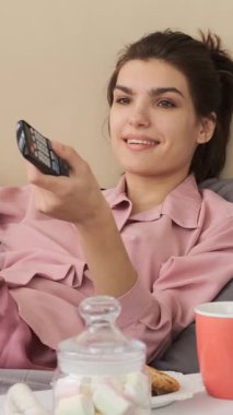 Pembe pijamalı mutlu bir genç kadın uzaktan kumandayla televizyon izler ve yatak odasında dinlenirken lezzetli bir kurabiye yer. dikey video