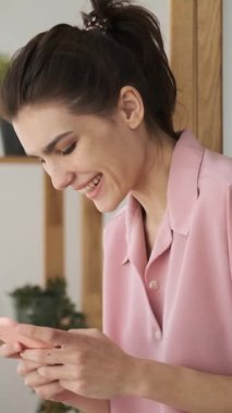 Pembe pijamalı neşeli genç bir kadın akıllı telefon kullanırken gülüyor ve gülümsüyor, komik bir mesajın ya da sosyal medya içeriğinin tadını çıkarıyor. dikey video