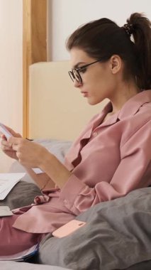 Gözlüklü ve pembe pijamalı genç bir kadın yatak odasında dizüstü bilgisayarla çalışırken iş belgelerini okuyor. dikey video