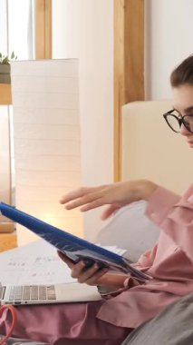 Pembe pijamalı odaklanmış genç bir kadın mavi bir klasördeki belgeleri inceler ve uzaktan çalışırken dizüstü bilgisayarında yazar. dikey video