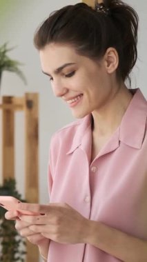 Pembe takım elbiseli mutlu genç bir kadın akıllı telefon kullanıyor. Sabahları yatak odasında dikilirken mesaj atıyor ve gülümsüyor. dikey video