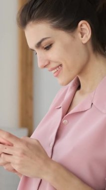 Pembe pijamalı neşeli genç bir kadın evde akıllı telefonuyla mesajlaşırken gülüyor ve gülümsüyor. İyi haberlerin ya da komik mesajların tadını çıkarıyor. dikey video