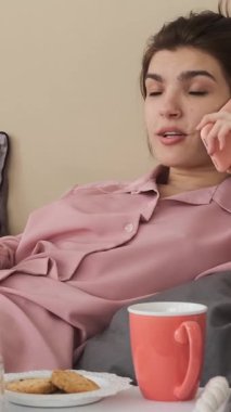 Pembe pijamalı mutlu genç bir kadın elinde bir televizyon kumandası ve kahvaltısıyla yatakta dinlenirken akıllı telefonuyla konuşuyor. dikey video