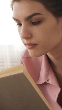 Genç bir kadın, evde pembe pijamalarıyla rahatlarken hikayeye gülümseyen bir kitap okuyor. dikey video