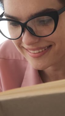 Siyahi gözlüklü genç bir kadının aşırı yakın plan çekimleri. İlgiyle kitap okuyor ve bir hikayeden zevk alırken gülümsüyor. dikey video