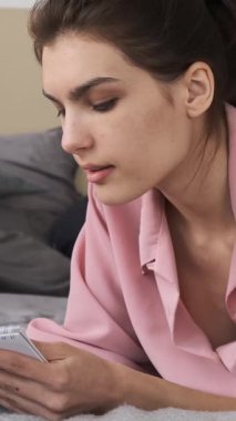 Pembe pijamalı mutlu genç bir kadın not defterine kalemle yazar, düşünür ve evinde yatağında yatarken gülümser. dikey video