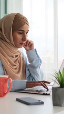 Laptop üzerinde çalışan ve belgeleri analiz eden tesettürlü genç Müslüman kadın. Dahili ofiste strateji düşünen ciddi bir serbest çalışan ya da öğrenci..